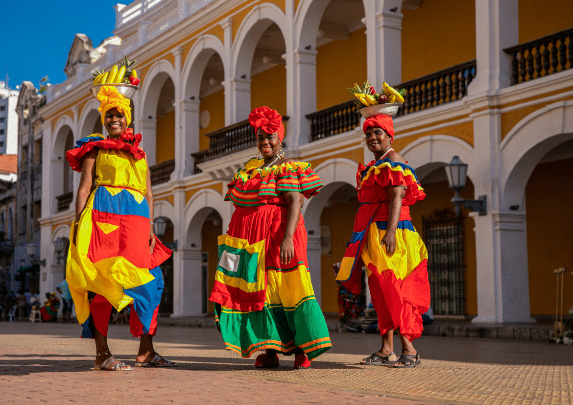 Trois femmes souriantes en robes traditionnelles colorées, portant des bols de fruits sur la tête, posant sur une place ensoleillée devant un bâtiment colonial jaune et blanc à Carthagène, Colombie.