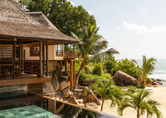 Villa luxueuse du Constance Lémuria Seychelles avec toit de chaume, piscine privée et vue sur la plage bordée de palmiers et de rochers granitiques typiques des Seychelles.