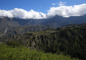 Paysage montagneux du Cirque de Cilaos à La Réunion, avec des reliefs verdoyants et des nuages accrochés aux sommets sous un ciel bleu.