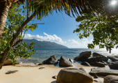Plage de sable doré bordée de rochers de granit et d’arbres tropicaux aux Seychelles, avec vue sur l’océan Indien et les montagnes verdoyantes.