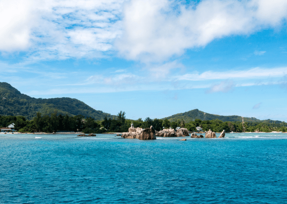 Vue depuis la mer sur l’île de La Digue aux Seychelles, avec ses rochers de granit typiques, ses collines verdoyantes et son lagon turquoise sous un ciel bleu éclatant.