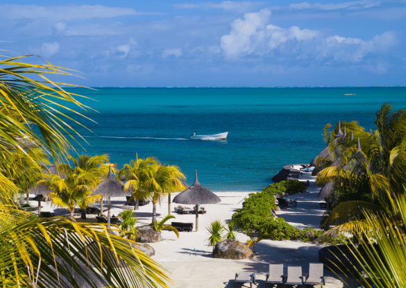 Vue sur une plage tropicale de l’île Maurice avec des palmiers, des transats, des paillotes et un bateau naviguant sur une mer turquoise.