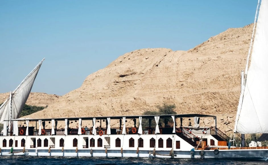 Dahabieh sur le Nil face aux falaises désertiques – Égypte Dahabieh naviguant sur le Nil avec de grandes voiles blanches et des falaises désertiques en arrière-plan.