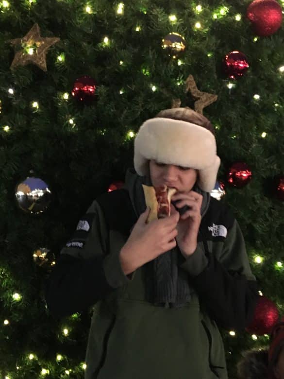 Personne dégustant un hot-dog devant un grand sapin illuminé sur un marché de Noël à Prague.