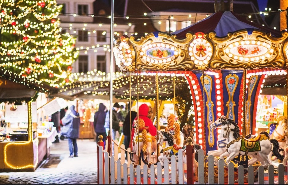 Marché de Noël européen illuminé avec un carrousel ancien, un grand sapin décoré et des visiteurs profitant de l’ambiance festive en soirée.