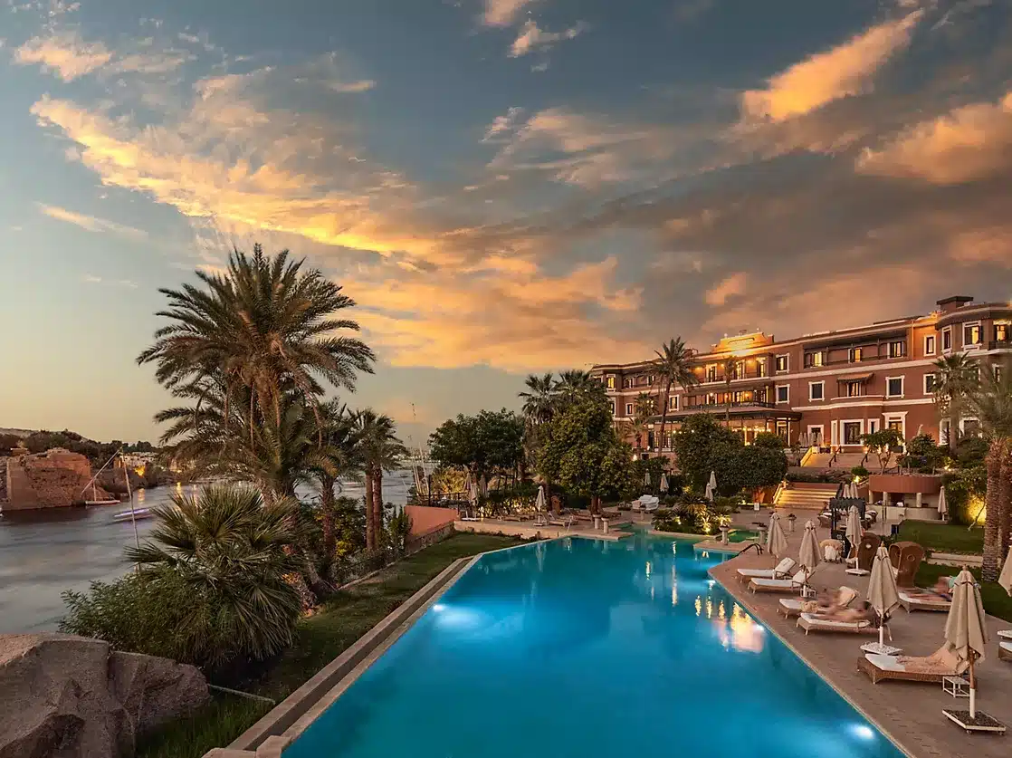 Sofitel Legend Old Cataract Aswan – Hôtel iconique au bord du Nil au coucher du soleil Vue du Sofitel Legend Old Cataract Aswan au bord du Nil, avec sa piscine et ses jardins illuminés par un coucher de soleil doré.