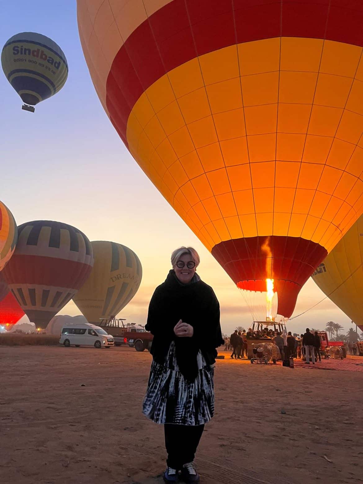 Vol en montgolfière au lever du soleil à Louxor – Égypte Personne debout devant une montgolfière en préparation au lever du soleil à Louxor, entourée de plusieurs ballons colorés prêts à décoller.
