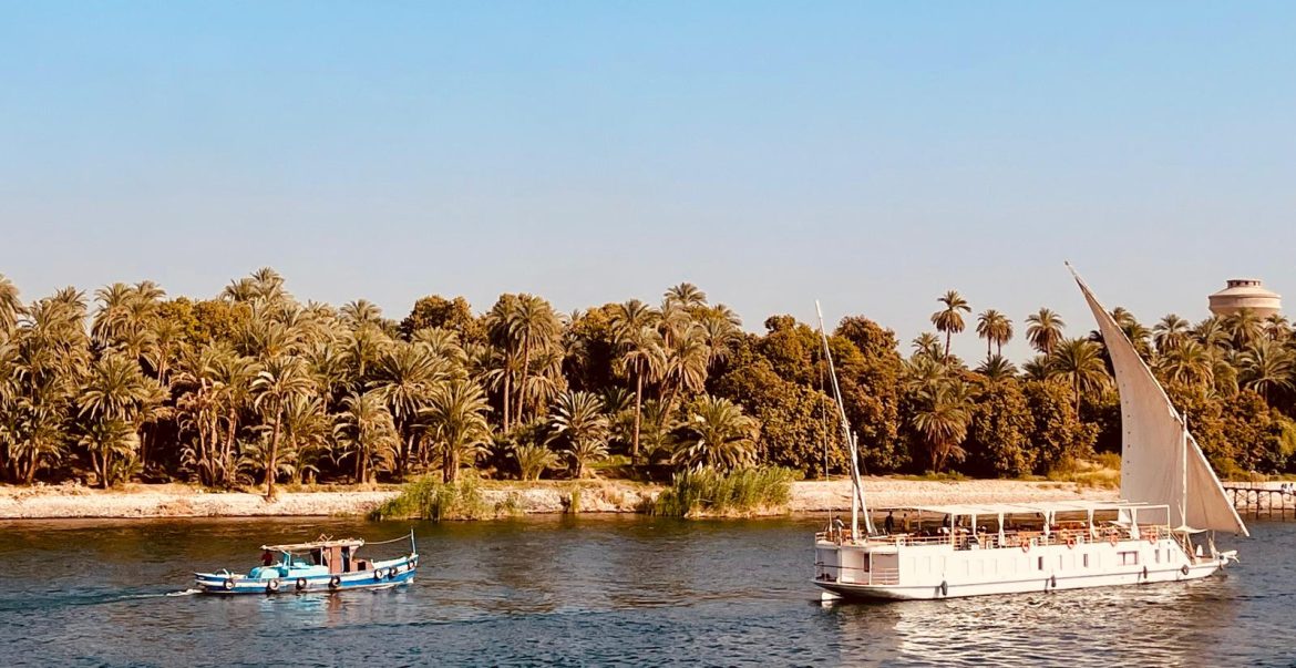 Deux bateaux naviguant sur le Nil, l’un à voile et l’autre motorisé, avec une berge couverte de palmiers sous un ciel bleu.