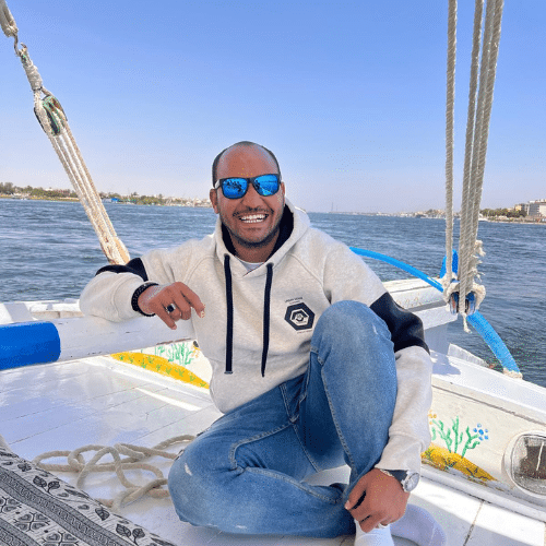 Guide souriant assis à bord d’une felouque naviguant sur le Nil par une journée ensoleillée.