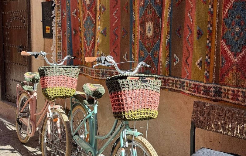 Deux vélos pastel avec paniers tissés, stationnés devant une façade décorée de tapis berbères au Maroc.